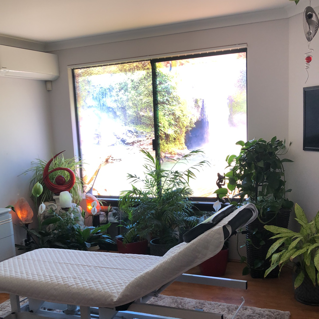 The treatment room in daylight, with a reclining massage table, a wall of pot plants and a waterfall print across the window.